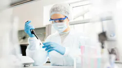 Serious concentrated female microbiologist in sterile clothing and safety goggles sitting at table and dropping reagent in petri dish while doing research in laboratory