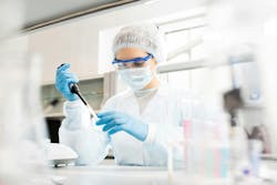 Serious concentrated female microbiologist in sterile clothing and safety goggles sitting at table and dropping reagent in petri dish while doing research in laboratory Serious concentrated female microbiologist in sterile clothing and safety goggles sitting at table and dropping reagent in petri dish while doing research in laboratory