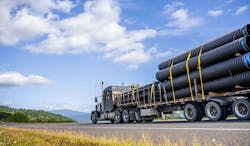 Big rig powerful professional industrial bonnet semi truck for long haul delivery commercial plastic pipes going with step down semi trailer on the summer road with green forest trees on the sides Big rig powerful professional industrial bonnet semi truck for long haul delivery commercial plastic pipes going with step down semi trailer on the summer road with green forest trees on the sides