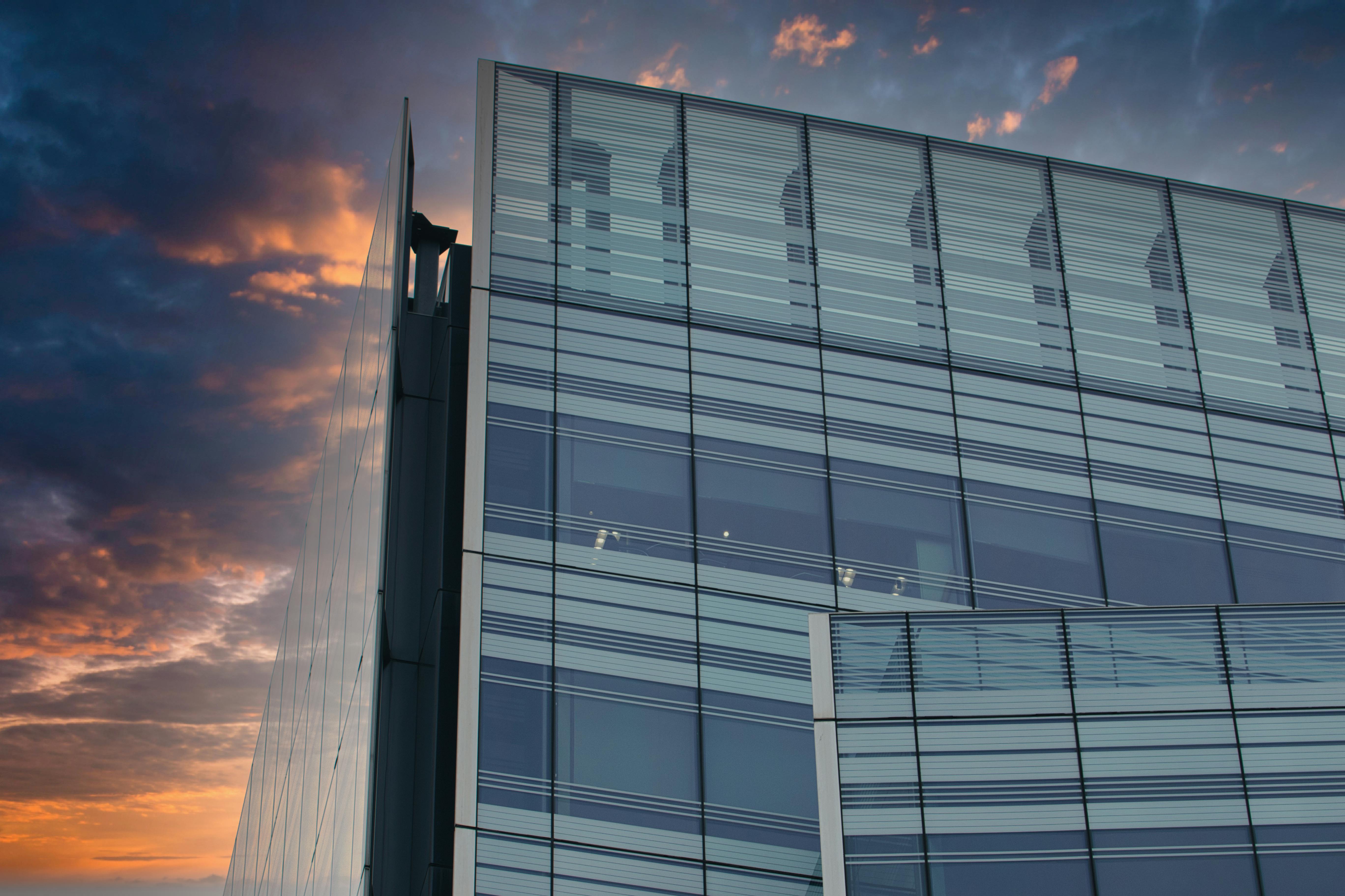A modern glass and steel building against a colorful sunset sky. The building s facade features large windows and a grid pattern, while the sky is a blend of orange, purple, and blue