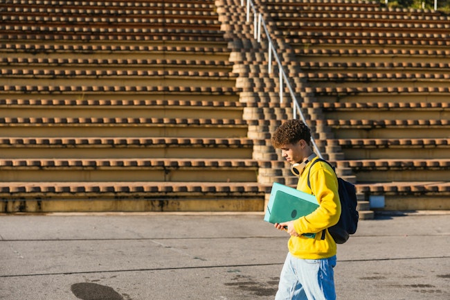 Young male college student walking near bleachers carrying laptop and backpack on sunny day