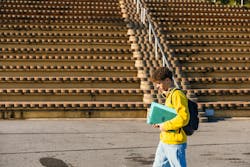 Young male college student walking near bleachers carrying laptop and backpack on sunny day Young male college student walking near bleachers carrying laptop and backpack on sunny day